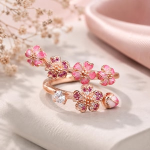May include: A rose gold bypass ring featuring multiple cherry blossom motifs. The flowers are accented with pink enamel, small round pink stones, and clear white stones. The band is open-ended, showcasing a delicate floral design with petals.