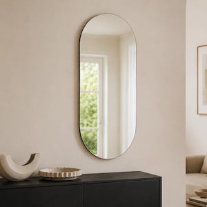 Op de afbeelding: Een hoge, ovaalvormige wandspiegel met een dunne, donkergekleurde lijst. De spiegel heeft een glad, reflecterend glazen oppervlak dat de omliggende binnenruimte, inclusief een raam en lichtgekleurde muren, in een verticale oriëntatie weergeeft.