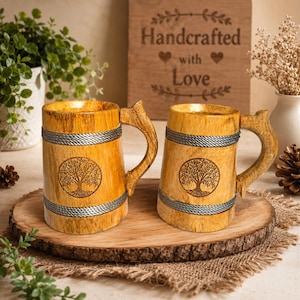Puede incluir: Dos jarras de madera de color marrón claro con veteado natural, cada una decorada con un grabado circular del árbol de la vida y dos bandas horizontales plateadas con diseño de cuerda. Las jarras tienen asas de madera resistentes.
