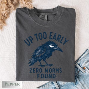 May include: A charcoal gray short-sleeve t-shirt featuring a dark blue graphic of a grumpy crow. The text above the crow reads UP TOO EARLY and the text below reads ZERO WORMS FOUND. The shirt has a crew neckline and a Comfort Colors brand label.