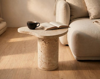 May include: A beige travertine side table with a cylindrical base and an irregular, organic-shaped top. A black ceramic mug filled with a dark liquid and an open book with white pages are placed on the surface of the stone table.