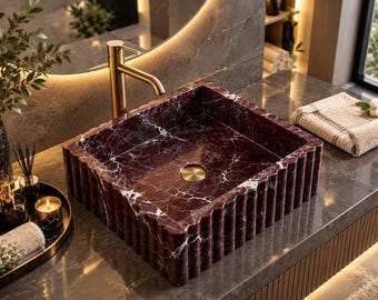 Pode incluir: Um lavatório quadrado feito de mármore vermelho profundo com veios brancos proeminentes. O exterior apresenta uma textura canelada vertical. Uma torneira de ouro escovado está montada atrás da bacia, que inclui um ralo circular a condizer.