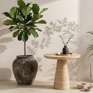 May include: A fiddle leaf fig plant in a large, dark, weathered ceramic pot stands next to a round, beige travertine stone side table. On the table sits a small, dark textured vase with dried stems on a gold tray, with two small stone bowls nearby.
