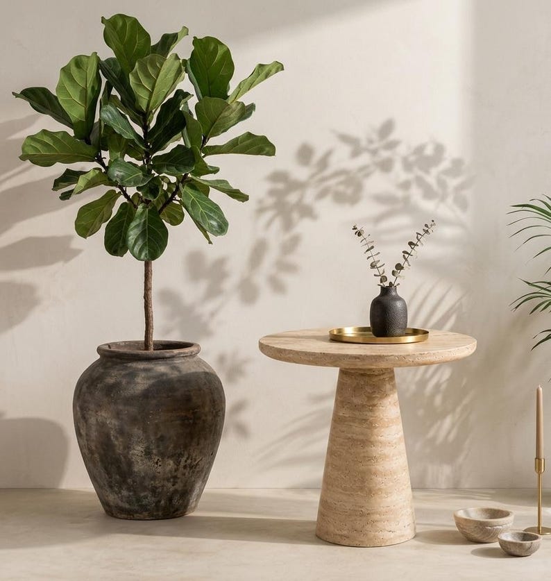 May include: A fiddle leaf fig plant in a large, dark, weathered ceramic pot stands next to a round, beige travertine stone side table. On the table sits a small, dark textured vase with dried stems on a gold tray, with two small stone bowls nearby.