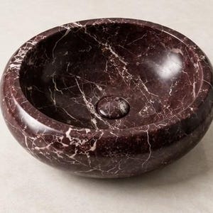 Pode incluir: Um lavat&oacute;rio redondo feito de m&aacute;rmore bordeaux profundo com veios brancos proeminentes por toda a pedra. A bacia apresenta uma superf&iacute;cie interior e exterior lisa e polida com uma tampa de drenagem circular a condizer no centro.