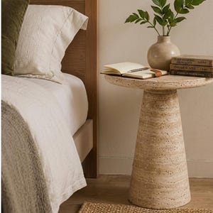May include: A beige travertine side table with a conical base and a circular top. On the table sits a light-colored ceramic vase with green leaves, an open notebook with a pen, and a stack of three books titled Sleep Medicine, Dream Scapes.