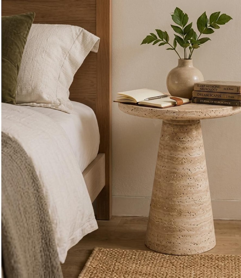 May include: A beige travertine side table with a conical base and a circular top. On the table sits a light-colored ceramic vase with green leaves, an open notebook with a pen, and a stack of three books titled Sleep Medicine, Dream Scapes.