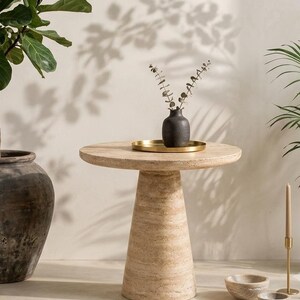 May include: A round beige travertine side table with a conical pedestal base. On the table sits a small black textured vase containing dried stems, placed on a circular gold tray. The table features a natural porous stone surface texture.