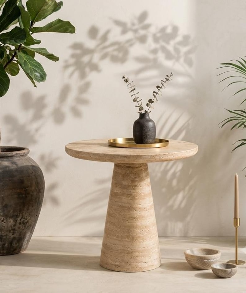 May include: A round beige travertine side table with a conical pedestal base. On the table sits a small black textured vase containing dried stems, placed on a circular gold tray. The table features a natural porous stone surface texture.