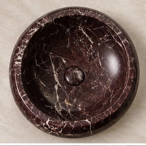 Pode incluir: Um lavat&oacute;rio circular de pousar feito de m&aacute;rmore bordeaux escuro com veios brancos proeminentes em toda a superf&iacute;cie da pedra. A bacia tem um acabamento liso e polido com uma tampa de ralo circular central que combina com o padr&atilde;o.