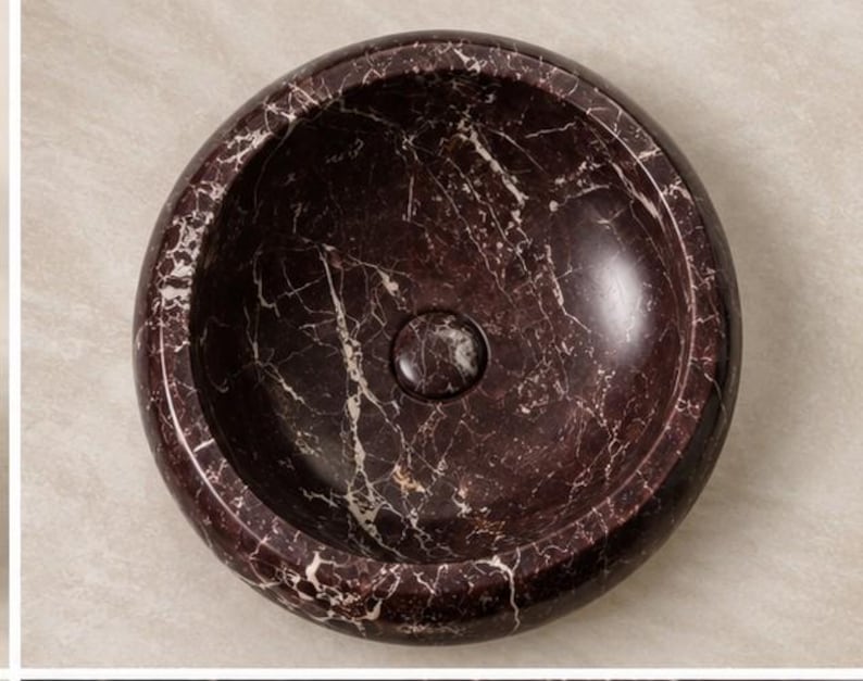 Pode incluir: Um lavat&oacute;rio circular de pousar feito de m&aacute;rmore bordeaux escuro com veios brancos proeminentes em toda a superf&iacute;cie da pedra. A bacia tem um acabamento liso e polido com uma tampa de ralo circular central que combina com o padr&atilde;o.