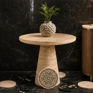 May include: A round beige travertine side table with a conical base, topped with a textured gray vase containing green foliage. Several circular coasters in black and beige with intricate mandala and floral patterns are arranged around the base.