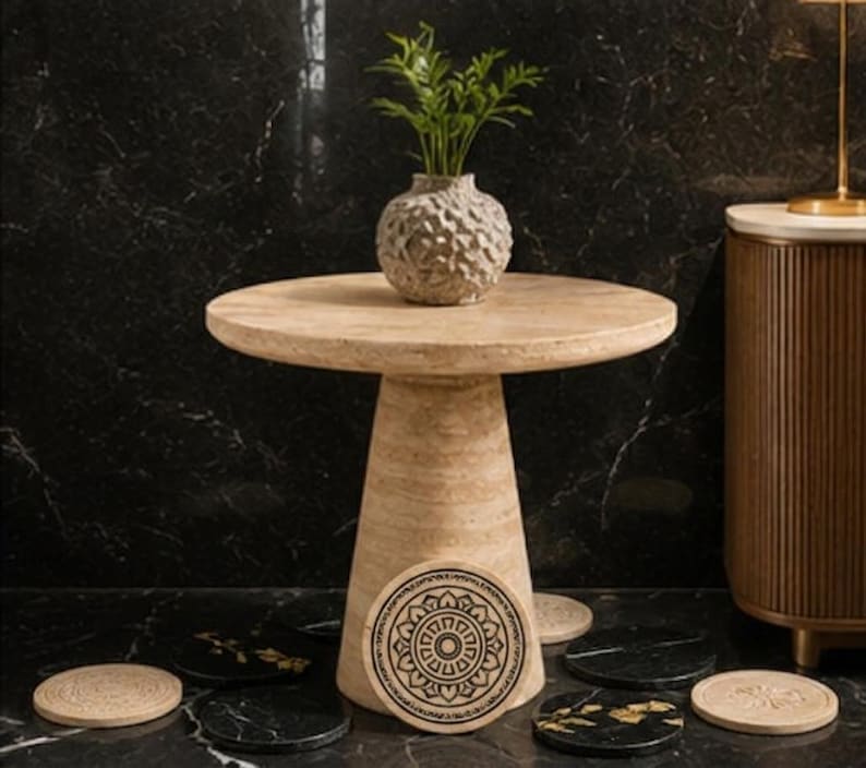 May include: A round beige travertine side table with a conical base, topped with a textured gray vase containing green foliage. Several circular coasters in black and beige with intricate mandala and floral patterns are arranged around the base.