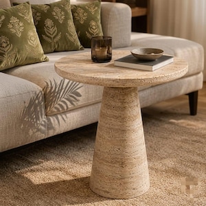 May include: A round beige travertine side table with a textured, porous surface and a tapered pedestal base. On the table sits a dark glass candle holder and a small, shallow bowl resting on a closed book with a white cover and dark spine.