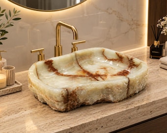 Pode incluir: Um lavatório retangular esculpido em pedra natural com interior polido em tons de creme e branco translúcido, acentuado por veios cor de ferrugem. O exterior mantém um acabamento bruto, texturizado e com uma forma irregular.