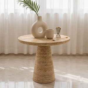 May include: A round beige travertine side table with a conical base. On the table sits a cream-colored donut-shaped ceramic vase with a green palm leaf, a small ribbed cylindrical beige cup, a clear glass jigger, and a thin gold-toned spoon.