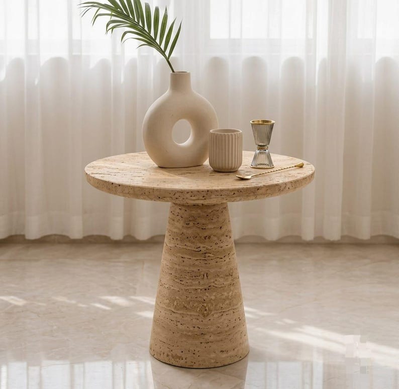 May include: A round beige travertine side table with a conical base. On the table sits a cream-colored donut-shaped ceramic vase with a green palm leaf, a small ribbed cylindrical beige cup, a clear glass jigger, and a thin gold-toned spoon.