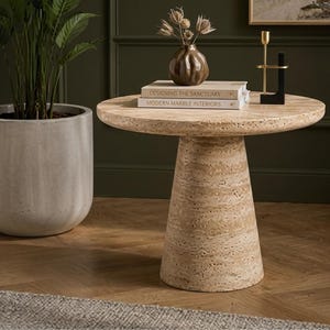 May include: A round beige travertine side table with a conical pedestal base. On the table sit two stacked books titled Designing the Sanctuary and Modern Marble Interiors, a small brown ceramic vase with dried flowers, and a gold and black candle holder.