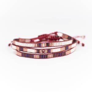 Puede incluir: Un brazalete de cuentas de triple vuelta con un cordón rojo y una mezcla de cuentas marrones, blancas y moradas.