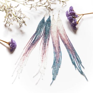 May include: Four pairs of long, beaded earrings in shades of white, pink, teal, and purple. The earrings are arranged on a white surface, with dried purple flowers in the background. The earrings have a gradient effect, transitioning from darker to lighter colors.