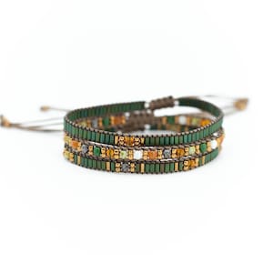 Puede incluir: Pulsera de cuentas de tres hilos con base verde, acentuada con cuentas doradas, naranjas y blancas. La pulsera tiene un diseño tejido y cierre de cordón ajustable. Las cuentas son pequeñas y rectangulares, creando un aspecto texturizado.