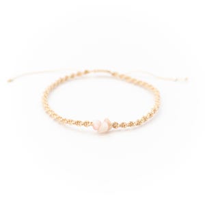 May include: A beige macramé bracelet with a small cluster of pink gemstones. The bracelet is knotted with a braided design and has adjustable ends. The gemstones are a soft pink colour, adding a delicate touch to the accessory.