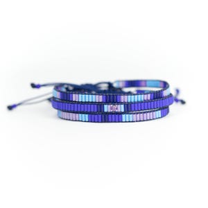 May include: Three layered beaded bracelets in shades of blue, purple, and light blue. The bracelets are tied together with a dark blue cord.