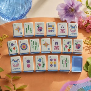 May include: A collection of rectangular mahjong tiles with white faces and blue translucent backs. The tiles feature colorful illustrations including ladybugs, flowers, joker faces, numbers, and Chinese characters, arranged in three rows on a surface.