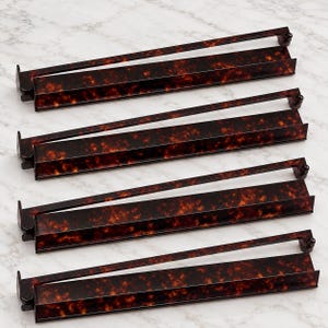 May include: A set of four rectangular hanger bars featuring a dark brown and black tortoiseshell pattern. Each bar has a flat base with a raised edge and a thin, horizontal top bar connected by a small circular hinge on one side.
