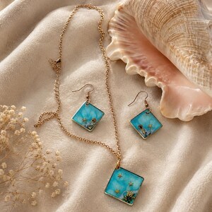 Puede incluir: Un collar y un par de pendientes a juego con colgantes cuadrados dorados. Cada colgante contiene una incrustación de resina azul brillante con pequeños dijes dorados en forma de estrella de mar y concha, suspendidos en cadenas finas.