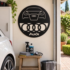 May include: A circular black metal wall sign featuring a cutout design of the front of an Audi car, the four-ring Audi logo, and the word Audi in white text. The sign is mounted on a wall above a wooden stool holding car cleaning supplies.