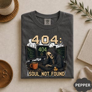 May include: A charcoal gray t-shirt featuring a graphic of a skeleton in a hoodie sitting at a desk with three computer monitors displaying code and 404 error messages. The text reads 404: SOUL NOT FOUND, accompanied by a coffee mug and a rubber duck.
