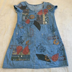 May include: A blue denim dress featuring a scoop neckline, short sleeves, and two front pockets. The fabric is decorated with various abstract shapes, including dark red teardrops, black geometric patches, and printed patterns in brown and black.
