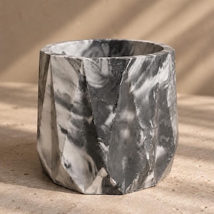 Peut inclure: Un pot de fleurs cylindrique doté d'un extérieur géométrique à facettes avec un motif marbré dans des tons de noir, de gris et de blanc. Le récipient présente une large ouverture circulaire sur le dessus et une base solide, avec une texture lisse.