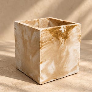 May include: A square-shaped planter with a marbled finish in shades of beige, cream, and tan. The container features a smooth, solid construction with sharp, defined edges and an open top, showcasing a swirling, organic pattern across its exterior.