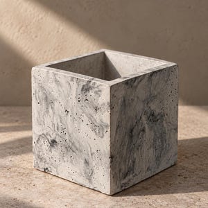 May include: A square-shaped planter made of gray and white marbled concrete. The exterior surface features a porous, textured finish with swirling dark gray and white patterns throughout the material, forming a hollow cube container.