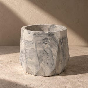 May include: A grey and white marble planter featuring a faceted geometric exterior with multiple flat surfaces. The stone vessel has a circular opening at the top and displays natural veining patterns throughout its solid, heavy construction.