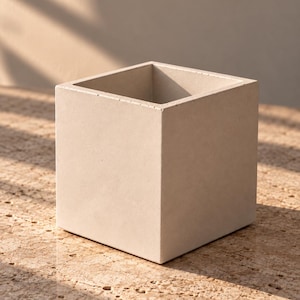 May include: A square-shaped planter with a smooth, light beige concrete finish. The container features a hollow center and flat, vertical sides, presenting a clean, geometric form with sharp edges and a uniform, matte texture throughout the exterior.