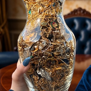 Pode incluir: Um vaso de vidro transparente cheio de uma grande coleção de joias em tom dourado, incluindo várias correntes, anéis, pingentes e pequenos berloques. O vaso é transparente, exibindo a densa variedade de itens metálicos, com o texto Maryam Heritage visível no canto superior direito.