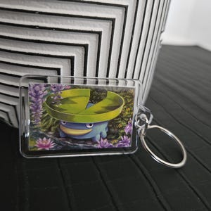 Può includere: Un portachiavi rettangolare in acrilico trasparente con un'illustrazione stampata di una creatura blu con una grande foglia di ninfea verde sulla testa, circondata da fiori viola e fogliame verde, fissato a un anello in metallo argentato.