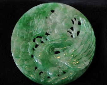 May include: A round, carved jade pendant with a phoenix design. The pendant is a vibrant green color and features intricate details of the bird's feathers and floral elements. The jade is polished and has a smooth surface.