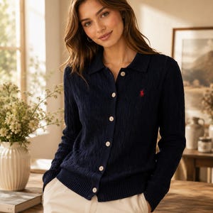 May include: A navy blue cable-knit cardigan featuring a collared neckline, a button-down front with light-colored buttons, and long sleeves. A small red embroidered logo is visible on the left chest area of the knit garment.