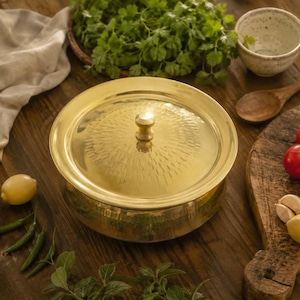 Puede incluir: Un plato de servicio redondo de latón pulido con una tapa a juego que presenta un pomo central y un patrón martillado texturizado en forma de rayos de sol en la superficie superior. El recipiente tiene un acabado liso y reflectante y un borde ancho.
