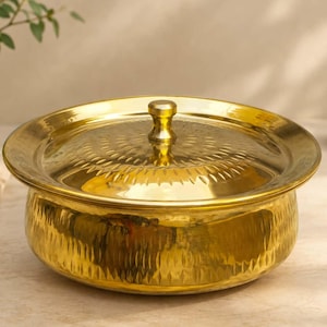 Puede incluir: Un cuenco de servicio redondo de metal dorado con tapa a juego. El exterior del recipiente presenta una textura martillada vertical, mientras que la tapa muestra un patrón radial de líneas grabadas y un pomo central para su manejo.