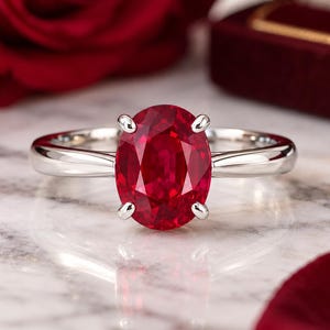 Peut inclure: Une bague argentée ornée d'une grande pierre précieuse rouge foncé de taille ovale, fixée par quatre griffes. L'anneau est lisse et poli, présentant un design épuré qui met en valeur la couleur vive et la surface facettée de la pierre.