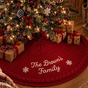 Op de afbeelding: Een ronde, donkerrode gebreide kerstboomrok met een dikke kabelgebreide rand. Het midden is geborduurd met de witte tekst "The Brown's Family", geflankeerd door twee witte sneeuwvlokpictogrammen, ontworpen om onder een versierde boom te liggen.