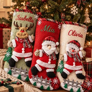 May include: Three knitted Christmas stockings in sage green, red, and cream, each featuring a plush character: a reindeer, Santa Claus, and a snowman. The stockings are personalised with the names Isabella, Mia, and Olivia, and decorated with plaid trim, white snowflakes, and a small fence detail.