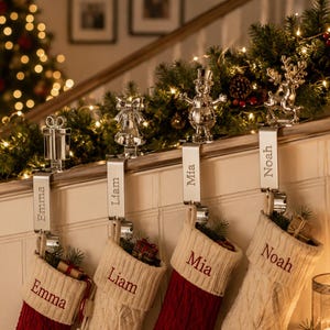 May include: Four silver-toned metal stocking holders with festive toppers including a gift, bell, snowman, and reindeer. Each holder features a vertical plate engraved with the names Emma, Liam, Mia, and Noah, holding matching knit stockings.