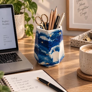 May include: A hexagonal desk tidy featuring a marbled blue and white pattern with small gold-coloured flecks embedded throughout. The container holds a pair of gold-coloured scissors, two wooden pencils, and two pens, standing on a wooden desk surface.