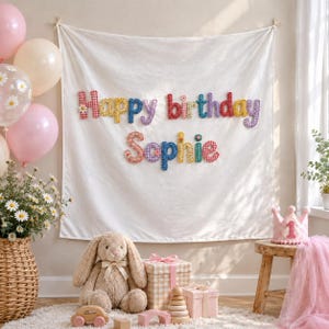 May include: A white fabric wall hanging featuring the colorful, fabric-stitched text Happy Birthday Sophie. The letters are patterned in various colors and textures, including gingham and floral prints, and are attached to the cloth backdrop.